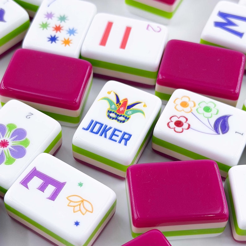 May include: A selection of Mahjong tiles with a variety of designs. The tiles have a white base with green and white stripes and magenta tops. Some tiles display floral patterns, numbers, and the word "JOKER." The game pieces are arranged closely together.