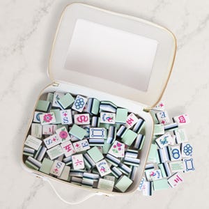 Puede incluir: Un estuche de transporte blanco con cremallera y espejo interior, lleno de una colecci&oacute;n de fichas de mahjong rectangulares. Las fichas tienen el reverso verde claro y el frente blanco con dise&ntilde;os impresos coloridos, incluyendo flores, n&uacute;meros y s&iacute;mbolos.