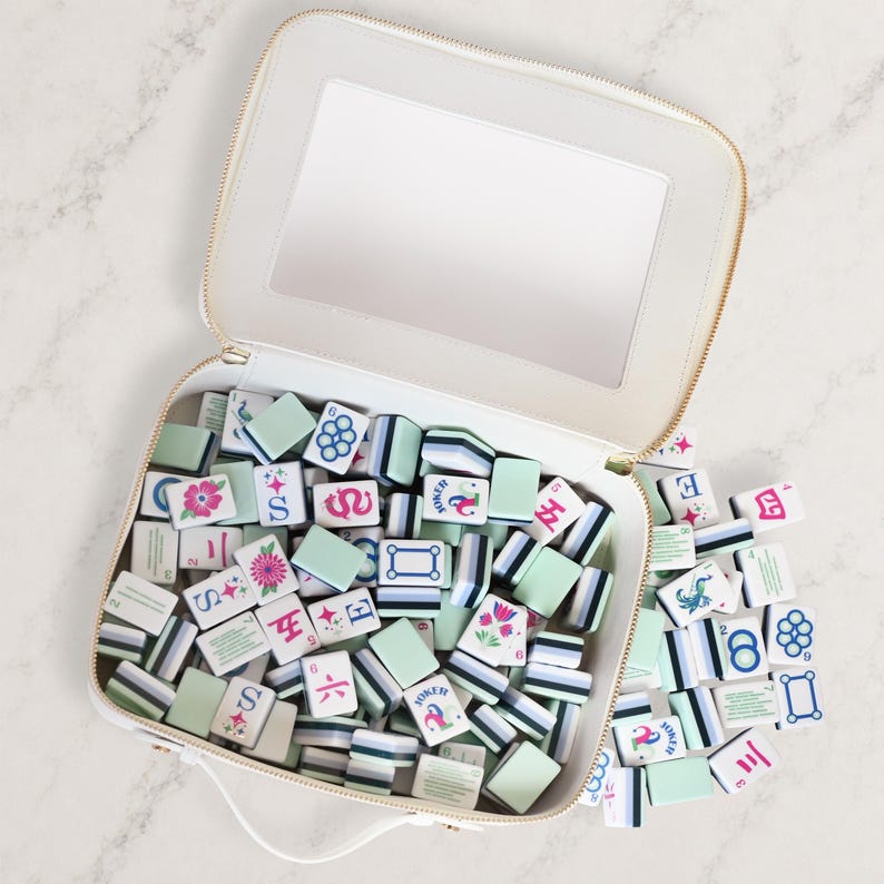 Puede incluir: Un estuche de transporte blanco con cremallera y espejo interior, lleno de una colecci&oacute;n de fichas de mahjong rectangulares. Las fichas tienen el reverso verde claro y el frente blanco con dise&ntilde;os impresos coloridos, incluyendo flores, n&uacute;meros y s&iacute;mbolos.
