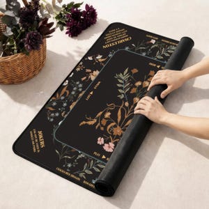 May include: A black rectangular game mat featuring floral illustrations in shades of orange, pink, and white. The mat includes printed text such as CHARLESTON, JOKERS, and various game instructions. The surface is smooth with a dark, matte finish.