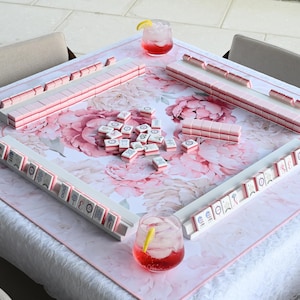 May include: A square mahjong table mat featuring a large pink and white floral peony pattern. The mat includes four white tile racks arranged in a square, holding pink and white mahjong tiles. A pile of loose tiles sits in the center with two glasses.