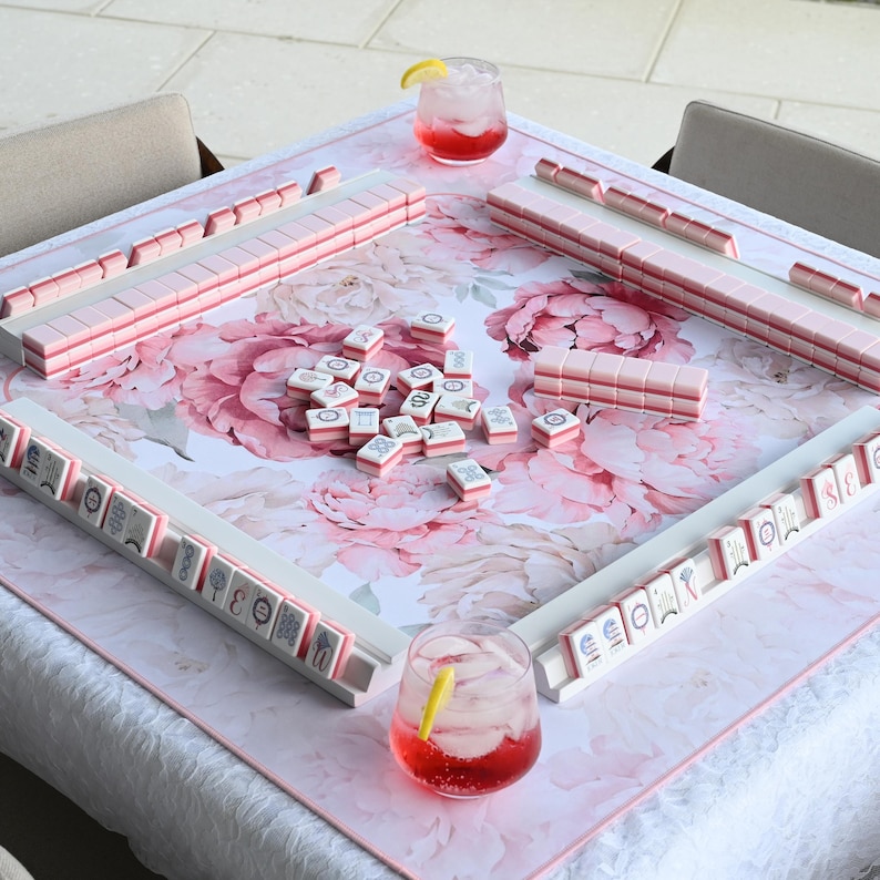 May include: A square mahjong table mat featuring a large pink and white floral peony pattern. The mat includes four white tile racks arranged in a square, holding pink and white mahjong tiles. A pile of loose tiles sits in the center with two glasses.