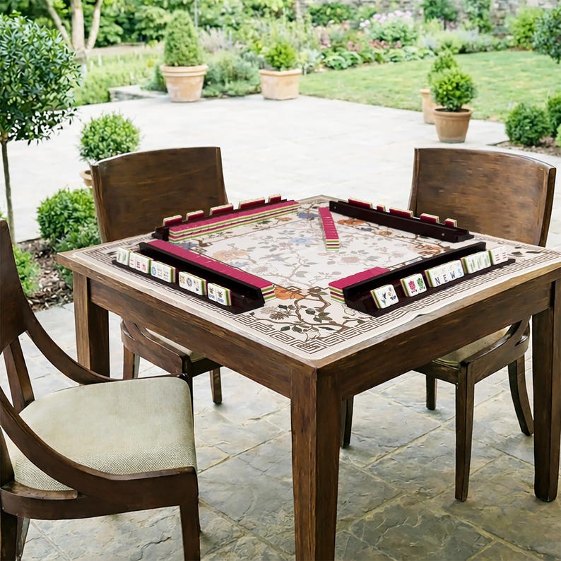 May include: A wooden mahjong table with a floral patterned top and four matching chairs. The mahjong set includes red tile racks and tiles. The scene is set outdoors on a patio with greenery in the background.