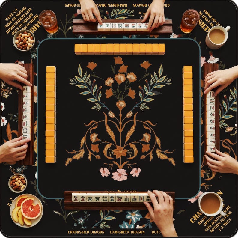 May include: A square mahjong mat with a black background featuring a central floral pattern in orange, pink, and green. The mat includes printed game instructions and is surrounded by four wooden tile racks holding ivory-colored mahjong tiles.