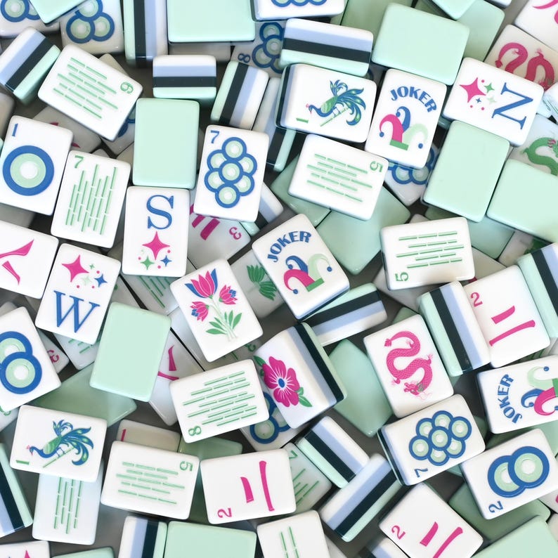 Puede incluir: Una colecci&oacute;n de fichas de mahjong rectangulares con reverso verde menta claro y caras blancas. Las fichas muestran varias ilustraciones coloridas, incluyendo flores, dragones, p&aacute;jaros, la palabra JOKER y patrones num&eacute;ricos en azul, rosa y verde.