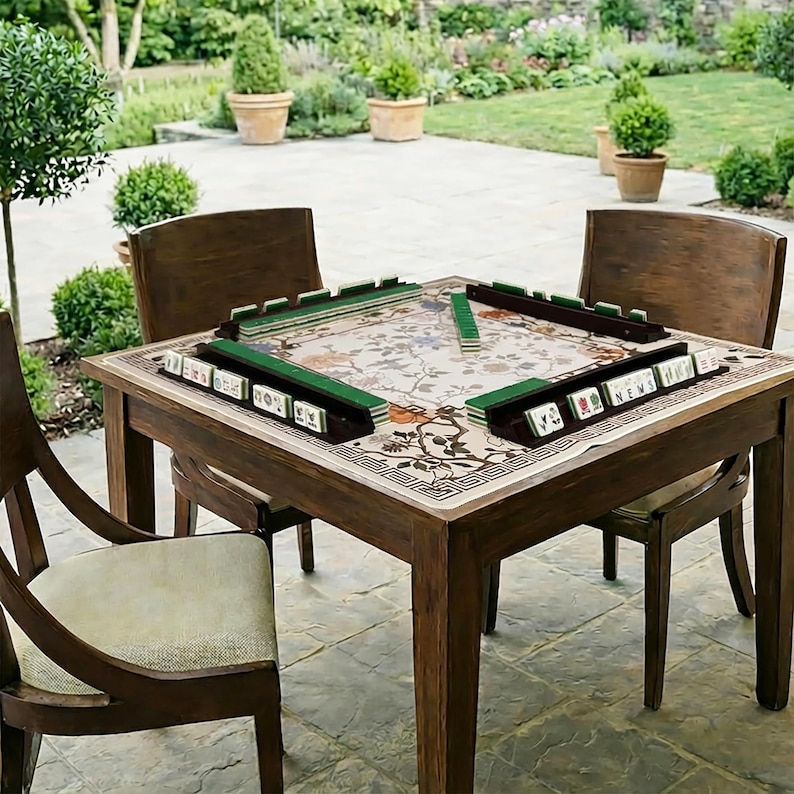 Puede incluir: Una mesa de mahjong de madera con una parte superior con estampado floral y cuatro sillas a juego. La mesa est&aacute; preparada con fichas verdes y blancas, lista para un juego. La escena es al aire libre, con un jard&iacute;n al fondo.