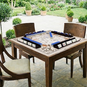 May include: A wooden mahjong table with four matching chairs is set up outdoors on a patio. The table has a floral patterned top with blue tile holders. The mahjong tiles are visible, ready for play. The chairs are made of dark wood.