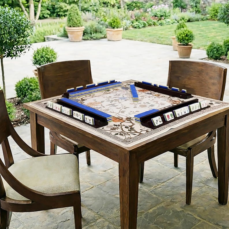 May include: A wooden mahjong table with four matching chairs is set up outdoors on a patio. The table has a floral patterned top with blue tile holders. The mahjong tiles are visible, ready for play. The chairs are made of dark wood.