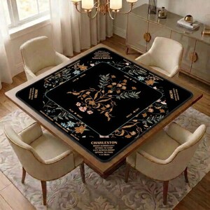 May include: A square black mahjong table mat featuring a central floral design in shades of gold, white, and pink. The border includes text reading Charleston, Jokers, and various game instructions printed in a clear, sans-serif white font.