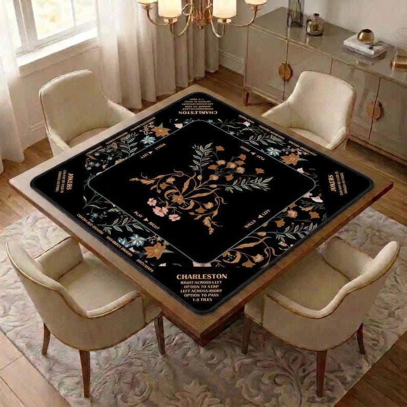 May include: A square black mahjong table mat featuring a central floral design in shades of gold, white, and pink. The border includes text reading Charleston, Jokers, and various game instructions printed in a clear, sans-serif white font.