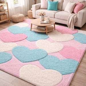 May include: A rectangular area rug with a pink background and large heart shapes in light blue and white. The rug is in a living room setting with a sofa, coffee table, and decorative items.