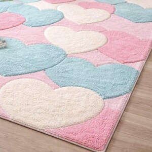 May include: A rectangular rug featuring a pattern of large hearts in pastel colors. The hearts are in shades of pink, light blue, and off-white, set against a pink background. The rug has a soft, plush texture.