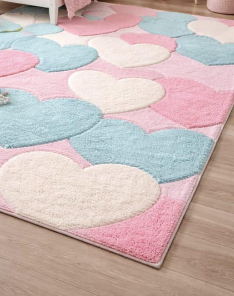 May include: A rectangular rug featuring a pattern of large hearts in pastel colors. The hearts are in shades of pink, light blue, and off-white, set against a pink background. The rug has a soft, plush texture.