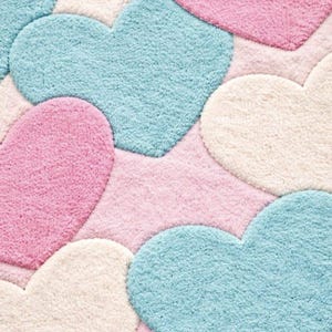 May include: A plush rug featuring a pattern of large, heart-shaped designs in pastel colors. The hearts are in shades of pink, blue, and cream, set against a pink background. The rug has a soft, textured appearance.