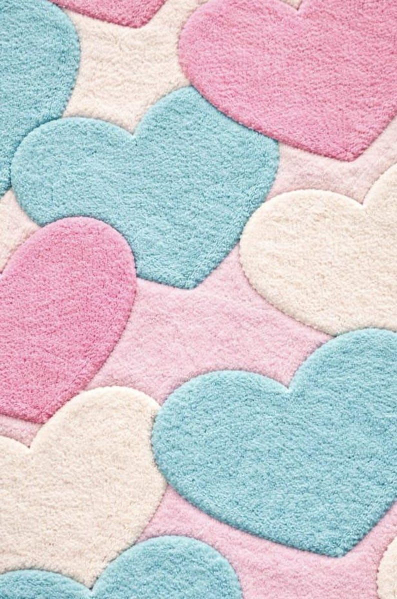 May include: A plush rug featuring a pattern of large, heart-shaped designs in pastel colors. The hearts are in shades of pink, blue, and cream, set against a pink background. The rug has a soft, textured appearance.