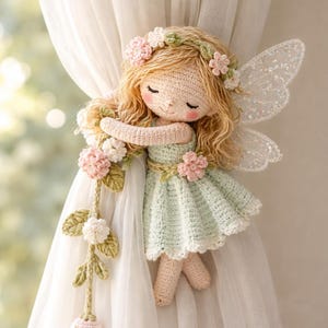 Könnte beinhalten: Eine handgefertigte Häkelfee-Puppe mit blonden Haaren, einem Blumenkranz und glitzernden Flügeln. Die Puppe trägt ein hellgrünes Kleid und hält eine Girlande aus rosa und weißen Blumen. Die Fee ist an einem weißen Vorhang befestigt.