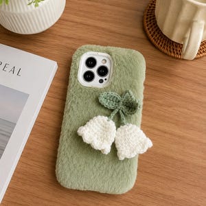 May include: A sage green fuzzy phone case featuring a white crocheted lily of the valley flower appliqué with two green leaves attached to the center. The case has a white camera cutout frame and a soft, plush texture covering the entire back surface.