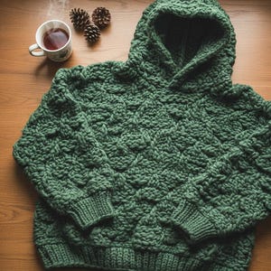 May include: A forest green, hand-knitted hoodie with a textured pattern. The hoodie features a large hood, ribbed cuffs, and a thick, cozy knit. A cup of tea and pinecones are next to the hoodie on a wooden surface.