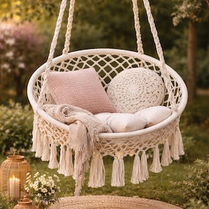 Op de afbeelding: Een crèmekleurige macramé hangstoel met een geweven ontwerp, kwasten en touwsteunen. De stoel is voorzien van roze en crèmekleurige kussens en een deken. Een rond geweven vloerkleed en decoratieve lantaarns met kaarsen staan op de voorgrond.