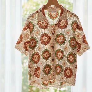 Puede incluir: Una camisa de crochet color crema con mangas cortas y botones. Presenta un patrón floral repetido en tonos naranja y verde. Cuelga de una percha de madera frente a una ventana.