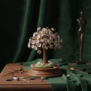 Puede incluir: Un árbol en miniatura artesanal con flores rosas y hojas verdes, colocado sobre una base de madera. La escena incluye una escultura de bronce, una pequeña botella y una cuchara dorada. El fondo es una cortina verde oscuro.