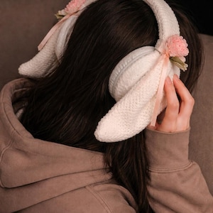 Può includere: Paraorecchie bianche lavorate a maglia con design a orecchie di coniglio. I paraorecchie presentano accenti floreali rosa e nastri. La persona nell'immagine indossa una felpa con cappuccio marrone.