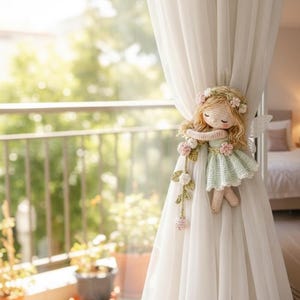 May include: A crocheted fairy doll with blonde hair and a floral crown, wearing a mint green dress, attached to a sheer white curtain. The fairy has small wings and is holding a garland of pink flowers. The background shows a balcony with plants.