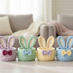 May include: Four pastel-coloured crocheted Easter baskets. Each basket, in lavender, green, yellow, and blue, features bunny ears and a bow tie. The baskets are displayed on a wooden surface.