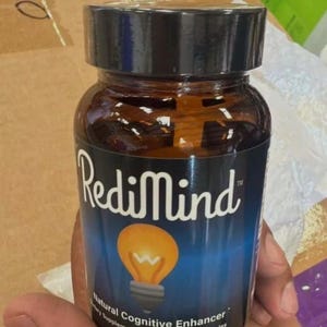 May include: A brown glass bottle with a black lid, labeled "RediMind." The label features a lightbulb graphic and text indicating it's a "Natural Cognitive Enhancer" with 30 vegetable capsules. The bottle is held in a hand.
