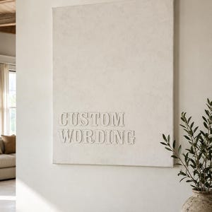 Puede incluir: Un lienzo rectangular de color blanco roto para pared que presenta el texto en relieve "CUSTOM WORDING" en una fuente con serifa. La superficie del lienzo tiene un acabado mate texturizado y las letras son del mismo tono monocromático.