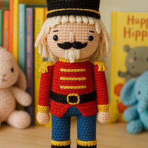 Könnte beinhalten: Gehäkelte Nussknacker-Puppe in einer roten Jacke mit goldenen Akzenten, blauen Hosen und schwarzen Stiefeln. Die Puppe hat einen schwarzen Hut mit goldenem Besatz, einen weißen Schnurrbart und Bart und steht aufrecht. Ein charmantes handgemachtes Spielzeug.