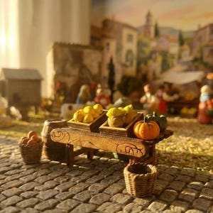 Peut inclure: Une scène de marché miniature avec une charrette en bois exposant des citrons, des courges et une petite citrouille. Des paniers d'oranges et un petit panier vide sont à proximité. La scène se déroule sur une rue pavée avec un arrière-plan flou de bâtiments.