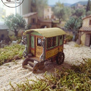 Op de afbeelding: Miniatuur houten caravan met een geel dak en groen en gele muren. De caravan heeft decoratieve krullen en een kleine deur met trappen. De scène speelt zich af op een zanderige ondergrond met miniatuurgebouwen op de achtergrond.
