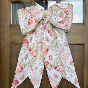 May include: A large, decorative bow made of quilted fabric, featuring a patchwork design with red roses and green accents. The bow is tied with a light green band and hangs on a wooden door. The bow's tails have pointed ends.