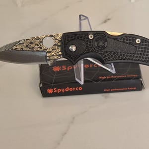 May include: A folding knife with a patterned Damascus steel blade featuring a circular thumb hole and a black textured handle with silver hardware. The knife rests on a black Spyderco box with white spiderweb graphics and the text High performance knives.