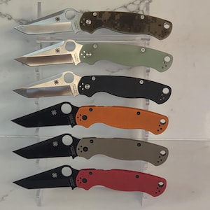 May include: A collection of six folding knives arranged vertically on a clear stand. The top three feature silver blades and handles in digital camouflage, olive green, and black. The bottom three have black blades with orange, tan, and red handles.