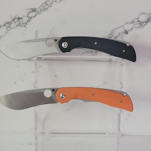 May include: Two folding knives with curved blades are displayed. The top knife has a black handle, while the bottom knife has an orange handle. Both knives have a silver blade and a circular hole near the blade's base.