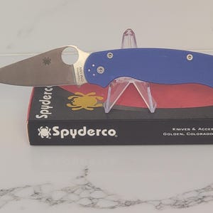 May include: A blue-handled folding knife with a silver blade, resting on a clear display stand. The knife is displayed on a black box with the brand name "Spyderco" and a yellow spider logo. The blade has a circular opening.