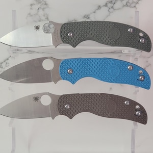 May include: Three folding knives with stainless steel blades and textured handles. The handles are in shades of green, blue, and brown. Each knife has a circular opening for one-handed deployment and a pocket clip.