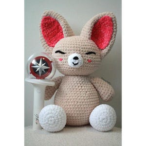 May include: A beige crochet plush toy with large ears featuring bright pink inner sections, closed eyes, and a white muzzle. Beside it stands a white light stick with a clear dome containing a silver star emblem and the text Stray Kids.