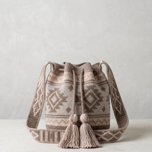 Könnte beinhalten: Eine beigefarbene und braune Häkel-Beuteltasche mit geometrischem Muster, einem Kordelzugverschluss mit zwei langen geflochtenen Schnüren, die in großen Quasten enden, und einem breiten, passenden gewebten Schulterriemen mit Rautenmuster.