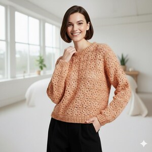 May include: A peach-colored, crew-neck sweater with a textured, open-weave design. The long-sleeved sweater has a ribbed hem and cuffs. The model is wearing black pants. The background includes a window, bed, and side table.
