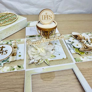 Może przedstawiać: Ręcznie robione pudełko o tematyce Pierwszej Komunii Świętej, otwarte, aby odsłonić elementy dekoracyjne. Pudełko zawiera motywy kwiatowe, kielich i litery "IHS". Zbudowane z jasnego drewna i papieru, ma delikatną, ręcznie robioną estetykę.