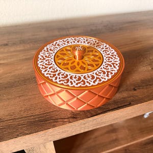May include: A round, orange decorative box with a lid. The lid features a gold center with a white, ornate design. The box has a diamond pattern on the sides and sits on a wooden surface. The box has a small knob on the top.