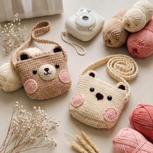 Puede incluir: Dos bolsos hechos a mano de ganchillo con forma de oso en beige y crema, cada uno con mejillas rosas y ojos negros. Los bolsos tienen correas largas y se exhiben con ovillos de lana y una cámara blanca.