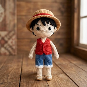 Può includere: Bambola all'uncinetto fatta a mano di un personaggio con un cappello di paglia con una fascia rossa, un gilet rosso, pantaloncini blu e scarpe beige. La bambola ha i capelli neri e un'espressione determinata. La bambola è su una superficie di legno.