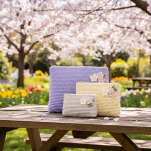 May include: Three crochet zipper pouches in lavender, yellow, and light gray, each adorned with floral embellishments. The pouches are displayed on a wooden picnic table, with a blurred background of a park with blooming trees.