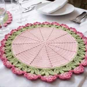 Könnte beinhalten: Ein handgefertigter, runder Häkel-Tischset in zartrosa Farbe mit einem grünen und rosa gewellten Rand. Das Tischset hat ein radiales Design, mit Linien, die vom Zentrum ausgehen. Das Tischset liegt auf einer weißen Tischdecke, mit anderen Gedecken sichtbar.