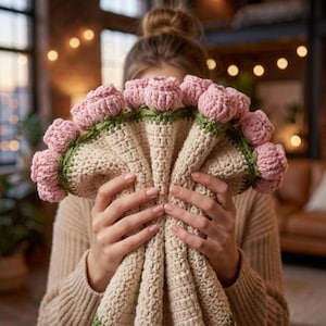 May include: A handmade crochet bouquet featuring pink flower buds with green stems and leaves. The bouquet is held in front of a person, with the flowers and stems arranged around a beige crocheted base. The background is blurred, with warm lighting.