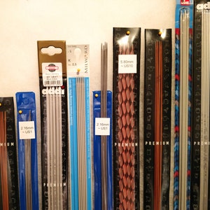 May include: A collection of knitting needles in various sizes and colors, including silver, copper, and wood. The needles are packaged in clear plastic and labeled with their size in millimeters and US size. The labels also include the brand names "Addi" and "Milward".